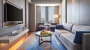 Une chambre d’hôtel moderne dotée d’une télévision à écran plat, d’un canapé, d’une table basse avec un panier de fruits et offrant une vue sur la ville grâce à de grandes fenêtres.