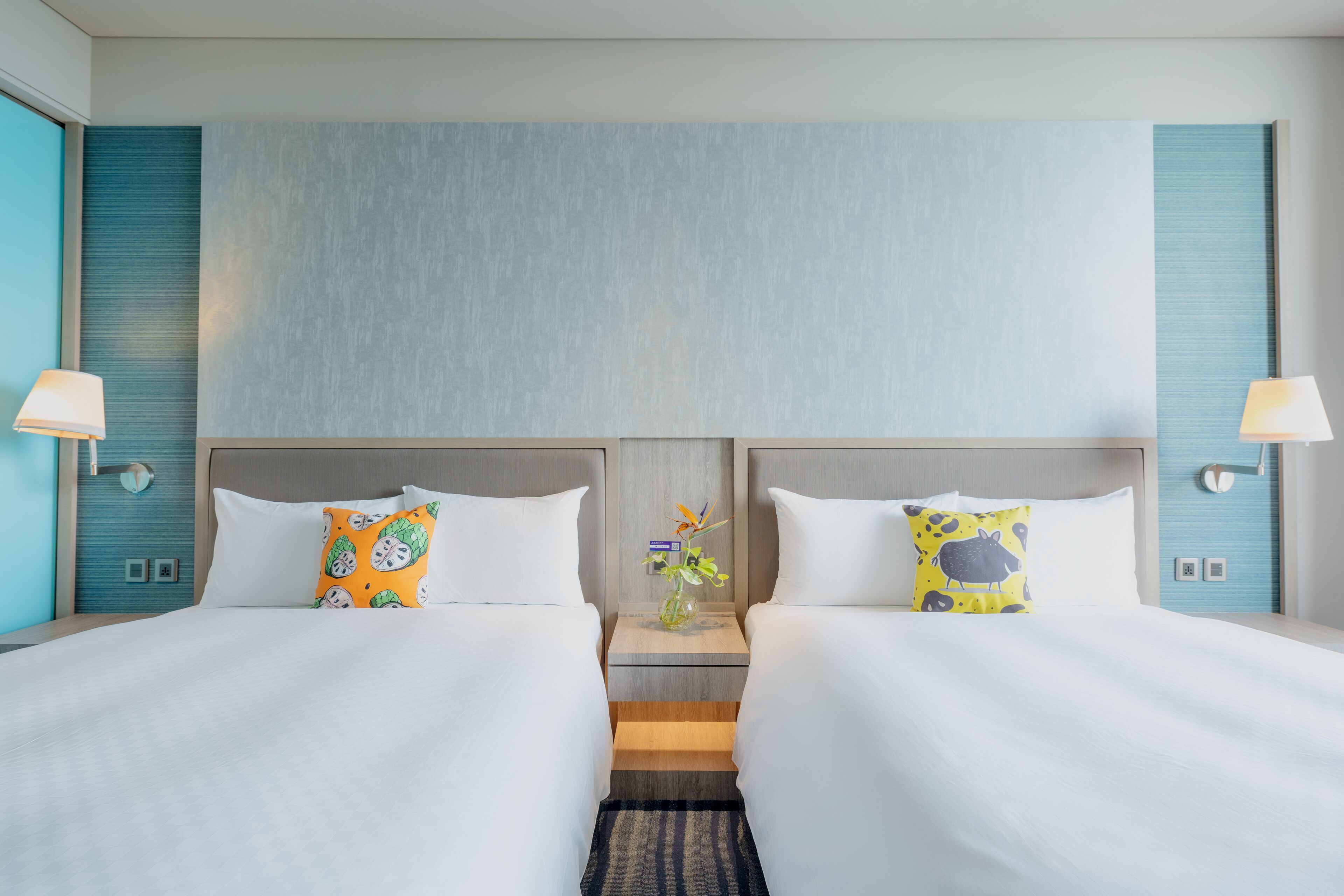 Habitación de hotel con dos camas, una mesita de noche con un jarrón de flores y lámparas fijadas a la pared.