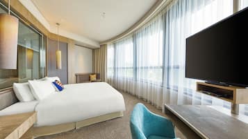 A modern hotel room with a large bed, a flat-screen TV, a blue armchair, and a large window with sheer curtains.