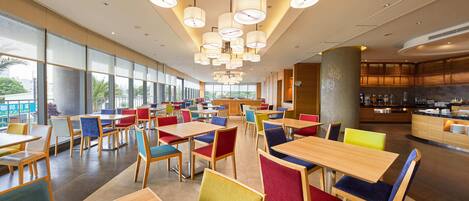 A spacious dining area with multiple tables and chairs, large windows, and a kitchen area in the background.