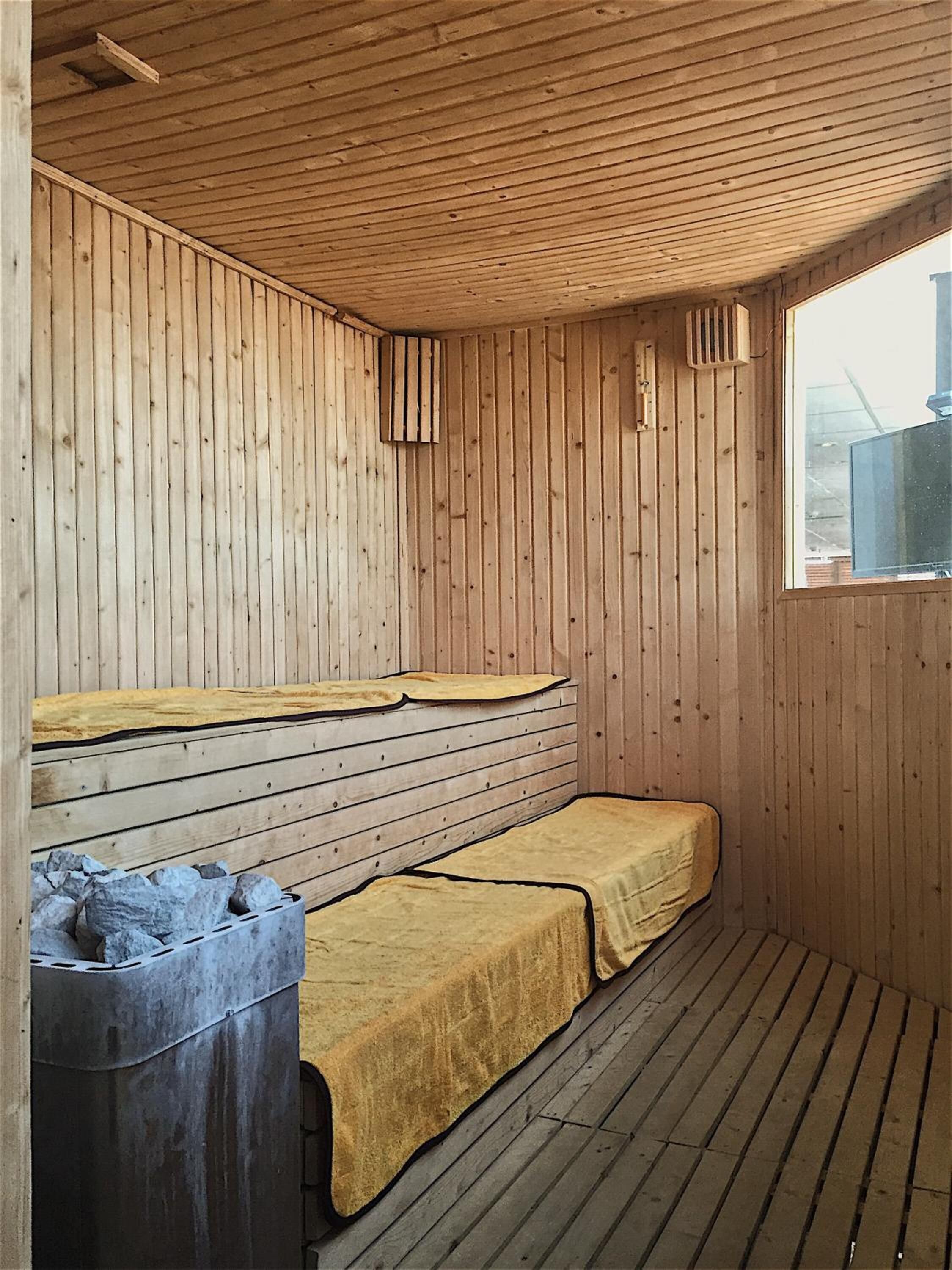 A sauna room with wooden walls and benches, a stone heater, and a window.