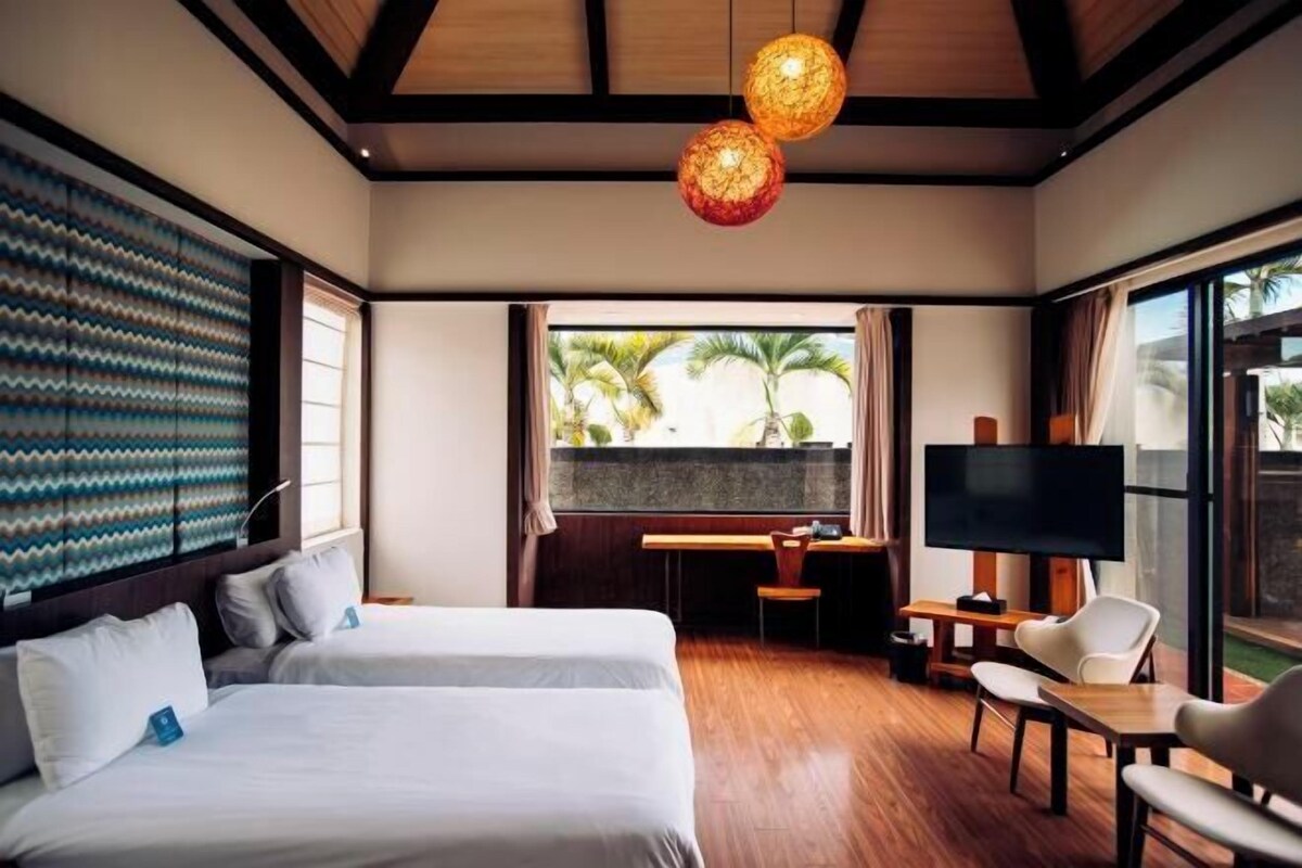 Une chambre d’hôtel équipée d’un lit, d’un bureau, d’une télévision et de grandes fenêtres donnant sur des palmiers.