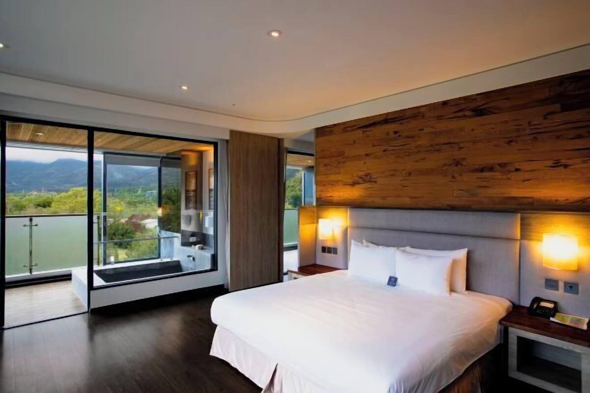 A modern hotel room with a large bed, a wooden accent wall, and a balcony with a view of the mountains.