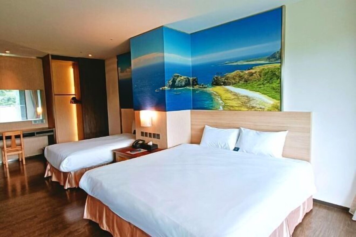 Une chambre d’hôtel avec deux lits, un bureau et un grand tableau représentant un paysage côtier.