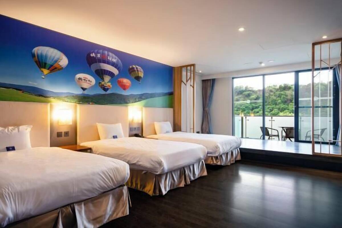 A hotel room with three beds, a mural of hot air balloons, a balcony with a view of greenery, and a large window.