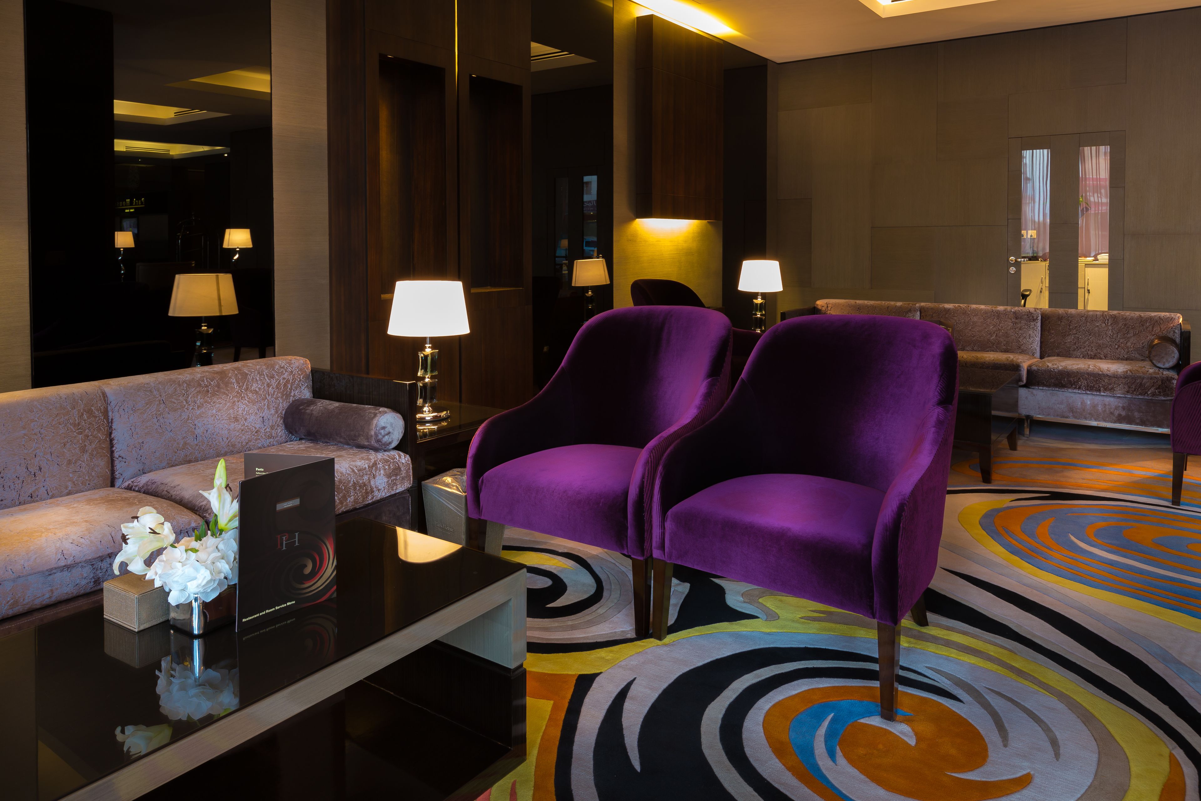 A modern hotel lobby with a sofa, two purple armchairs, a coffee table, and a colorful rug.