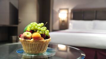 Una cesta de mimbre con fruta sobre una mesa de vidrio en una habitación de hotel, con una cama y una mesita de noche al fondo.