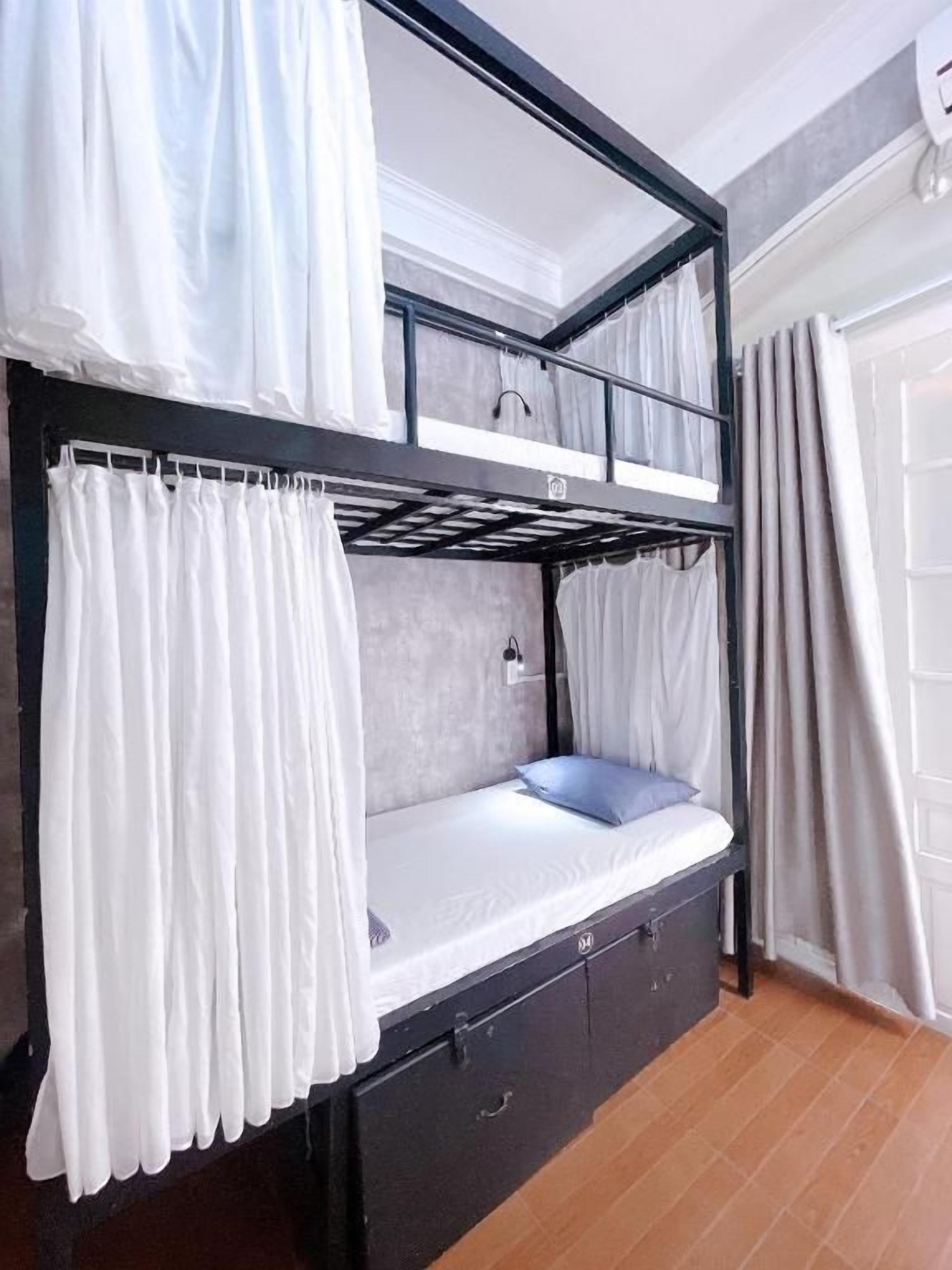 Una cama litera con cortinas blancas, un cajón pequeño y una ventana con cortinas.