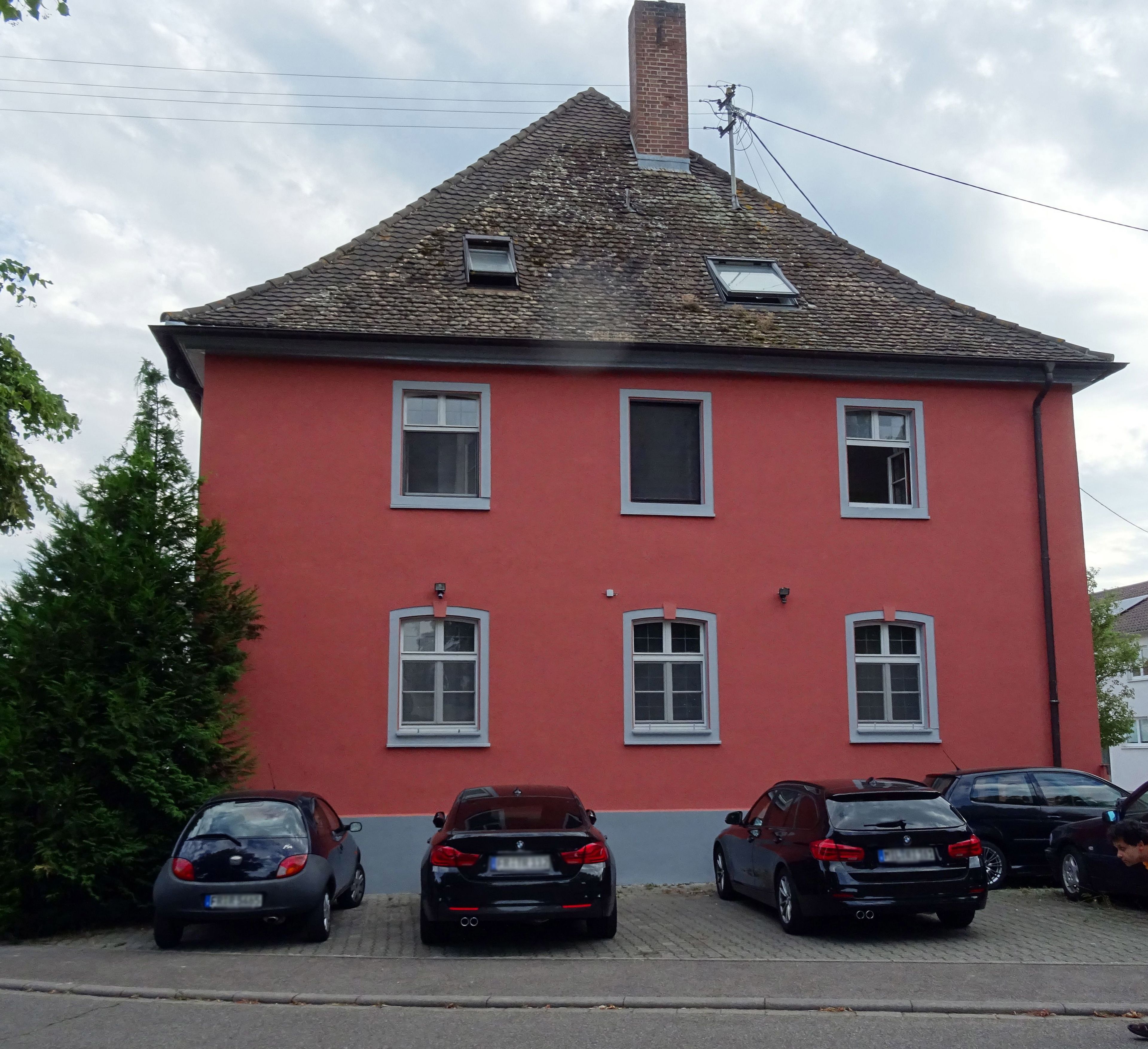 A red building with multiple windows and a sloped roof. There are several parked cars in front.