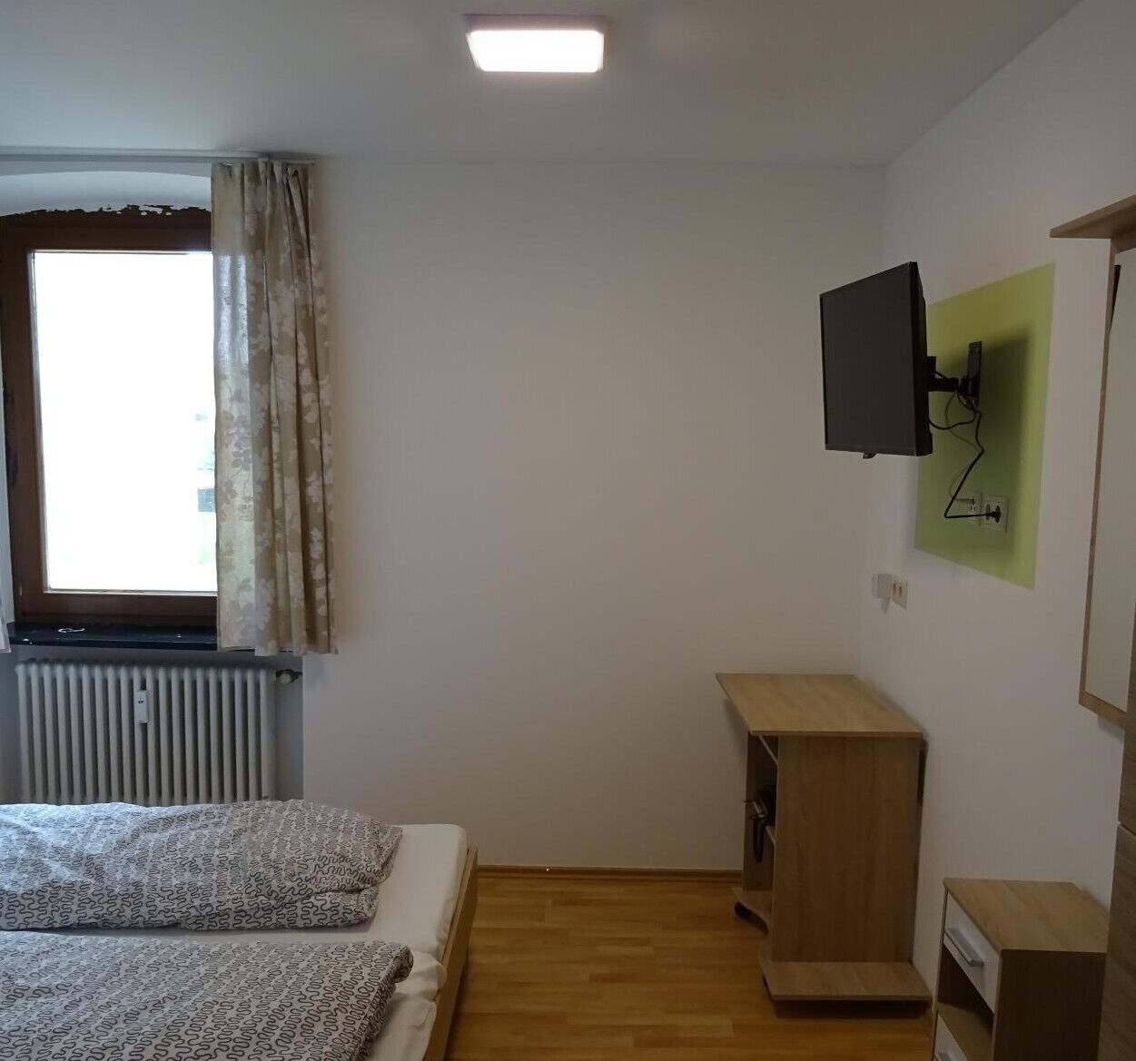 A small, sparsely furnished room with a bed, a TV mounted on the wall, and a radiator.