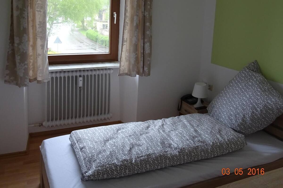A single bed with patterned bedding, a window with curtains, a bedside table with a lamp, and a radiator.