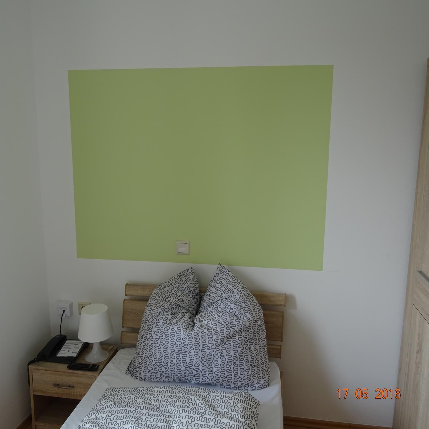 A bedroom with a bed, two patterned pillows, a bedside table with a lamp, a phone, and a wall-mounted light switch.