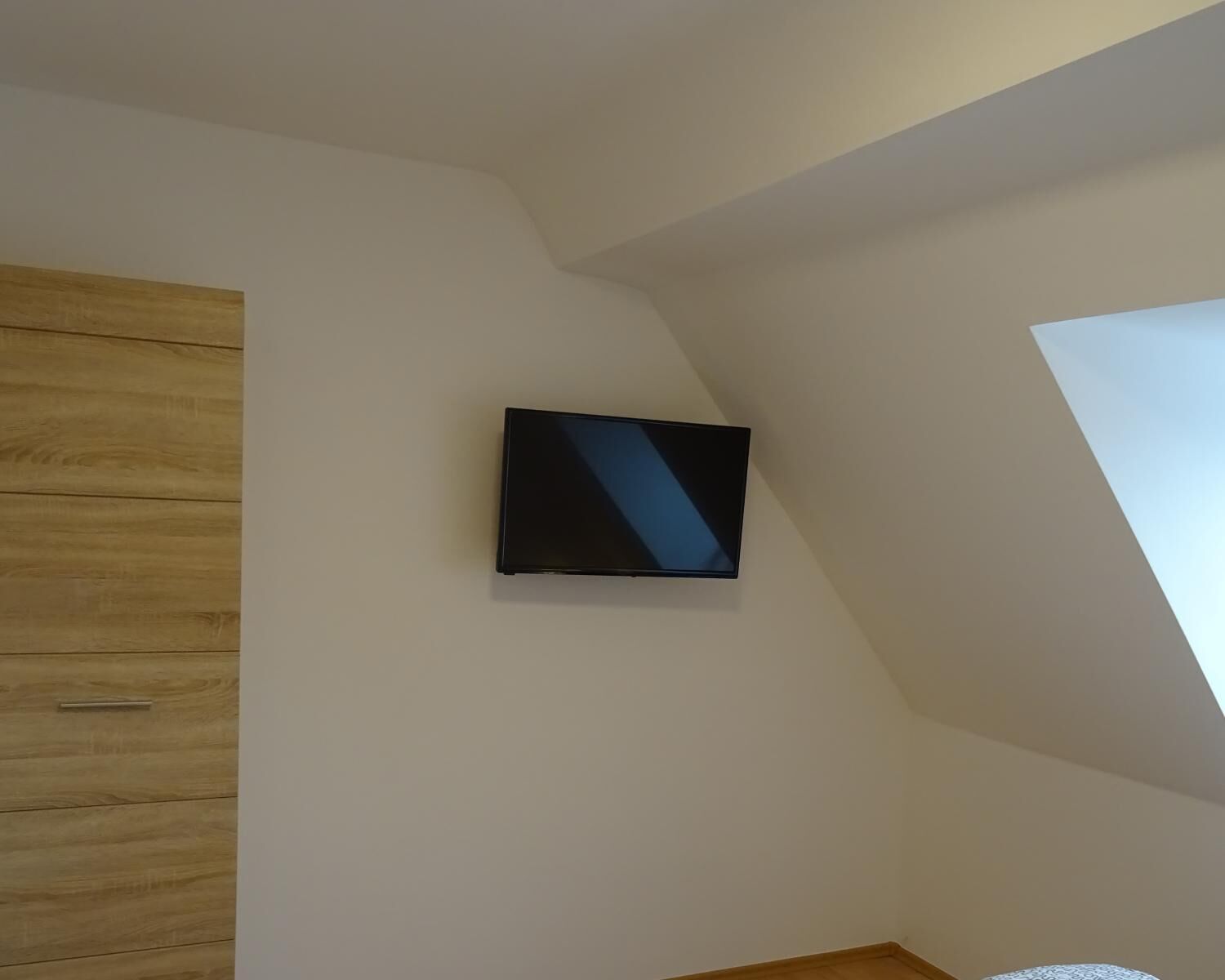 A room with a sloped ceiling, a wooden wardrobe, and a mounted television.