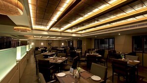 A spacious restaurant with multiple dining tables and chairs, a long counter with a lighted display, and a high ceiling with recessed lighting. - Goutham Grand Hotel (Tenali)