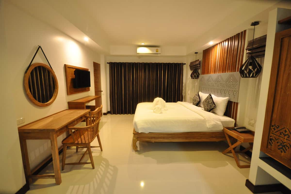 Habitación de hotel con una cama de madera, un escritorio y una silla. Tiene una ventana con cortinas, un aire acondicionado de pared y una pequeña mesa con lámpara.