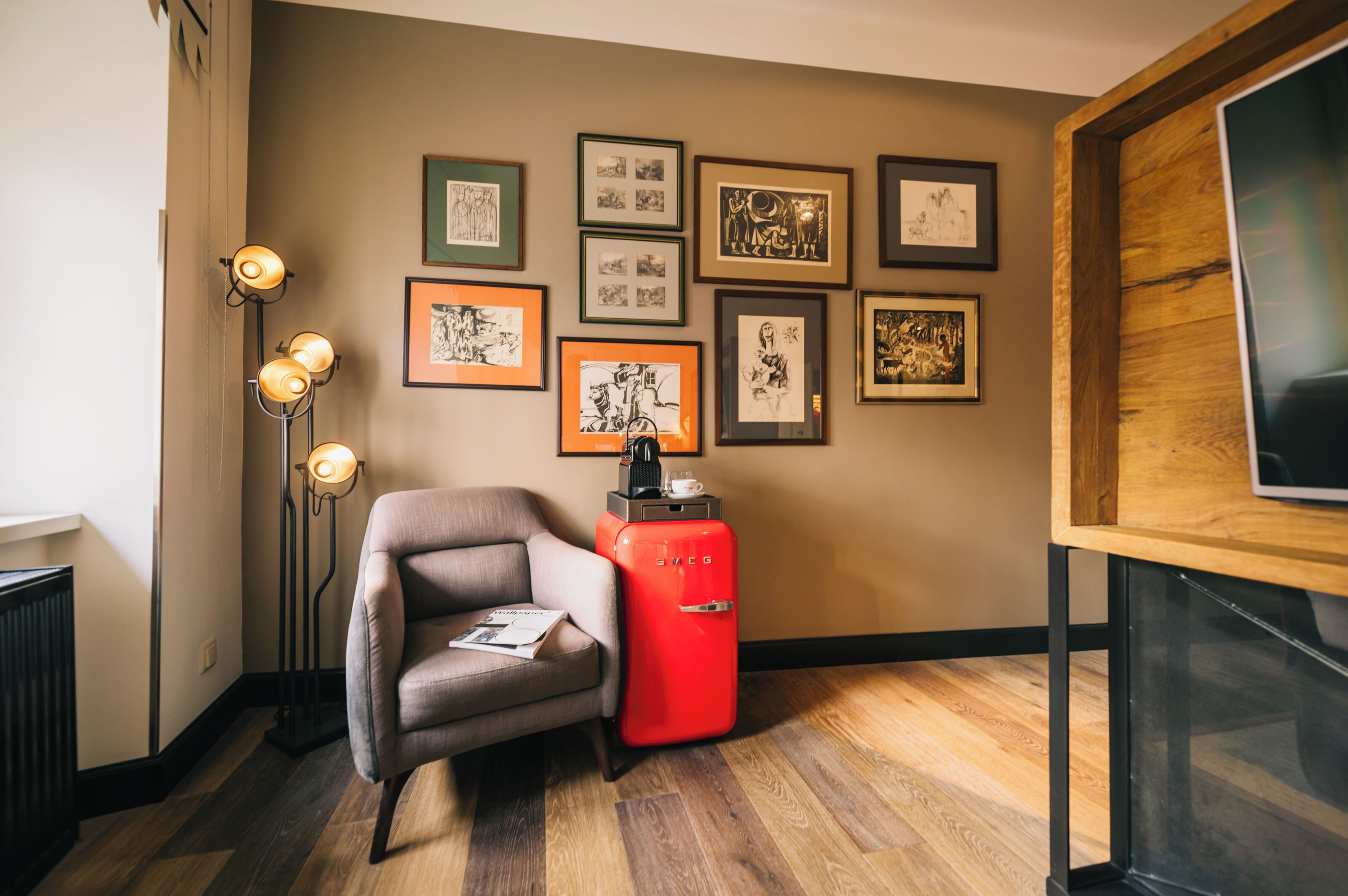 Una habitación con piso de madera, un refrigerador rojo de estilo vintage, un sillón gris, una lámpara de pie y un muro decorado con obras de arte enmarcadas.