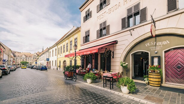 Una calle adoquinada con un hotel llamado PEST-BUDA. Hay mesas y sillas al aire libre, y una bicicleta estacionada.
