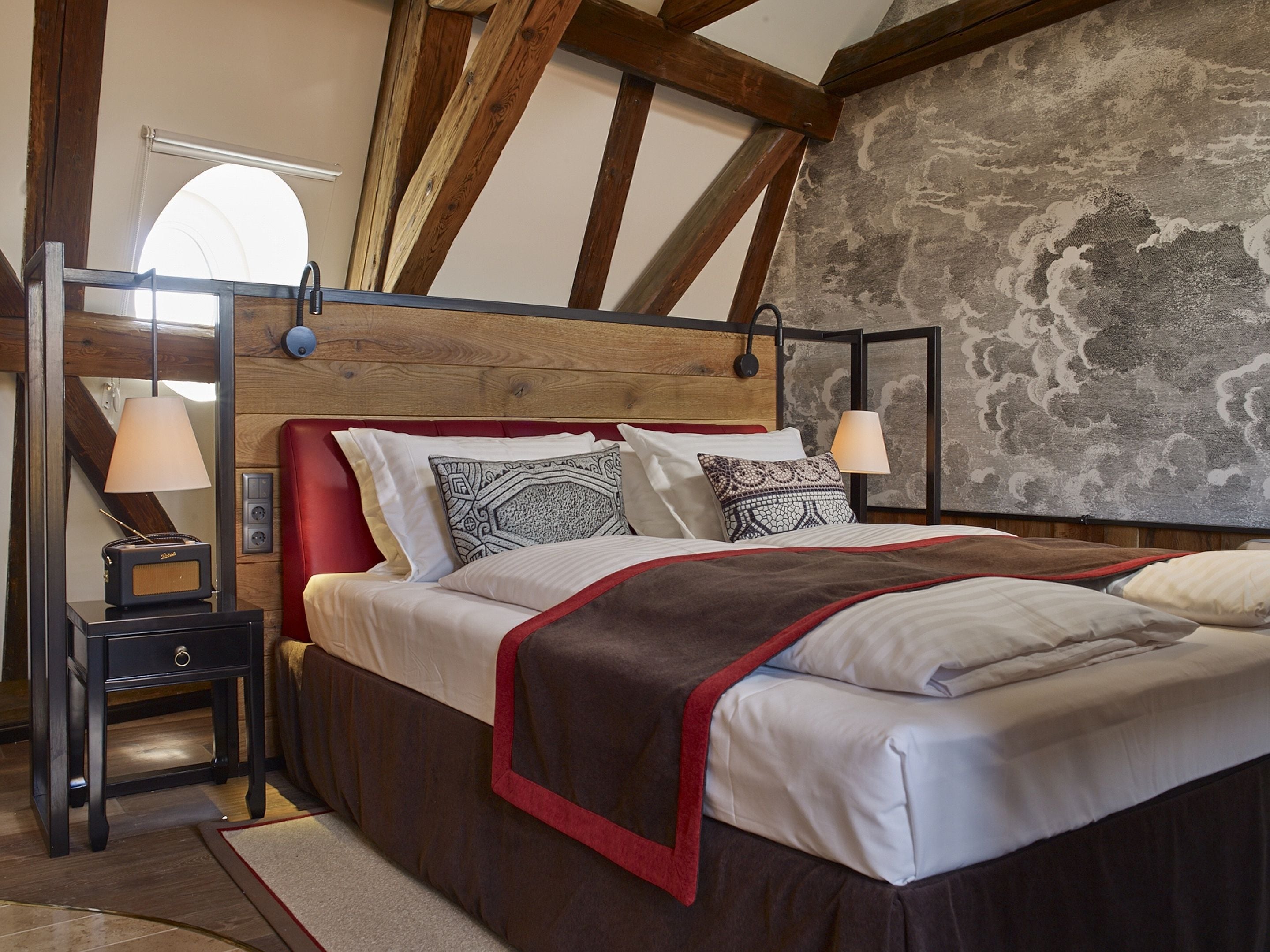 A bedroom with a wooden ceiling, a bed with patterned pillows, a nightstand with a lamp, and a stone wall.