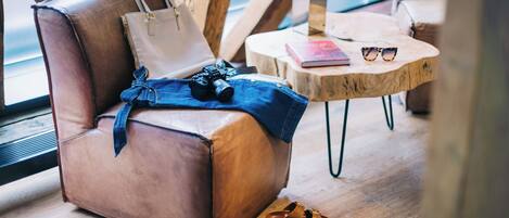 A leather chair with a camera, denim jacket, and sandals on the floor, a wooden table with a book and sunglasses, and a trophy on a wooden stool.