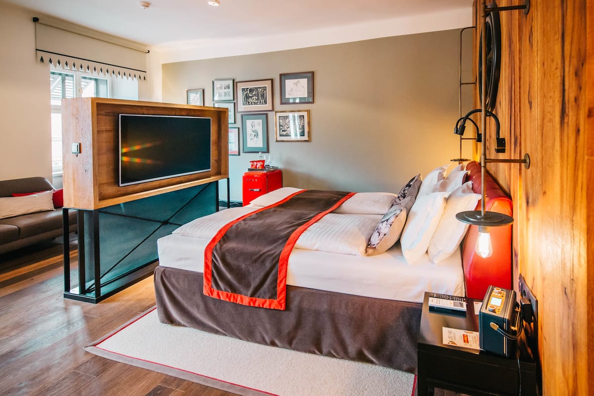 A modern hotel room with a large bed, a flat-screen TV, a red bedside table, and a wall-mounted TV.