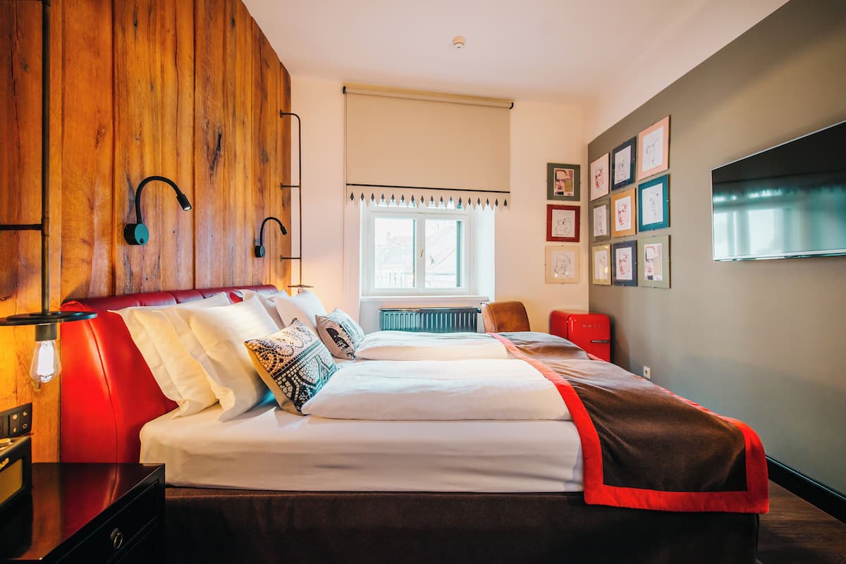 A bedroom with a wooden headboard, a bed with white bedding, a red accent wall, a window with a view, and a flat-screen TV mounted on the wall.