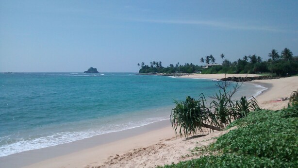A beach with clear turquoise water, a sandy shore, and a small island in the distance. - Jungleside House (Hikkaduwa)