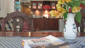 A dining table with a magazine, a vase with flowers, and a view of a kitchen with wooden cabinets and a red countertop. - Jungleside House (Hikkaduwa)
