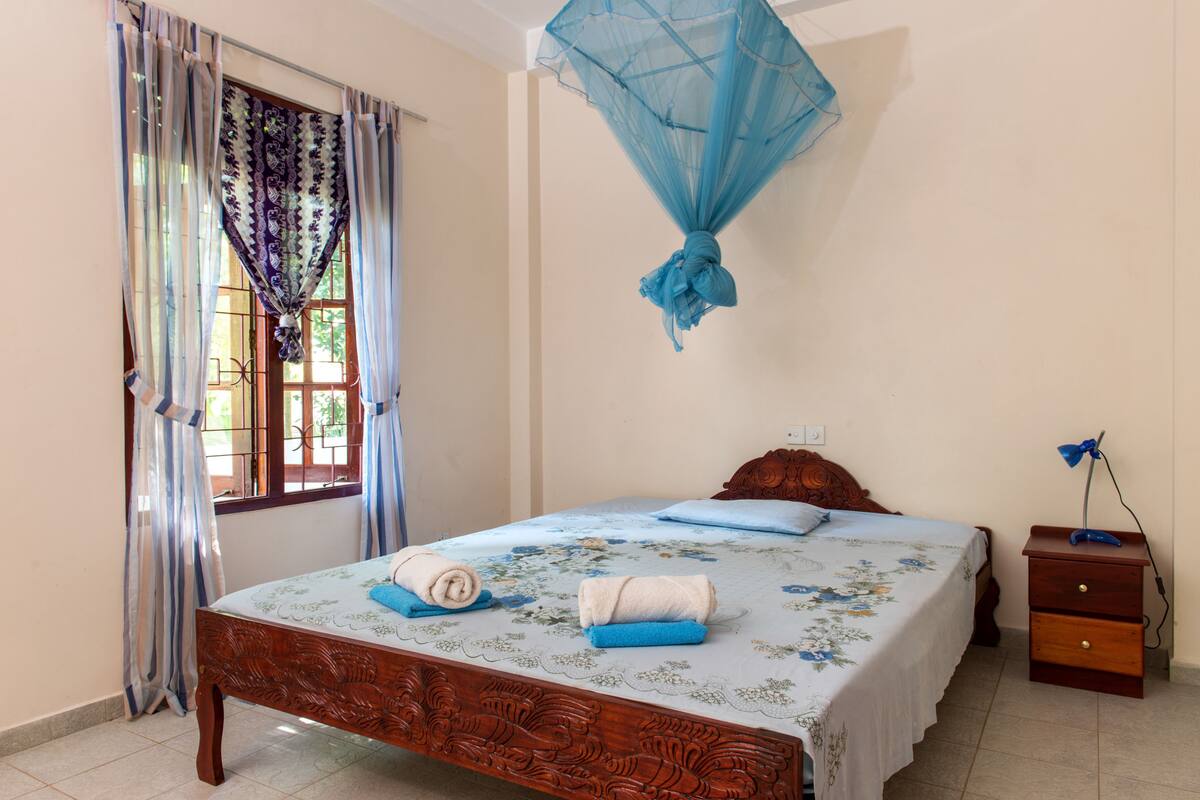 A bedroom with a bed, curtains, a mosquito net, a nightstand, and a lamp.