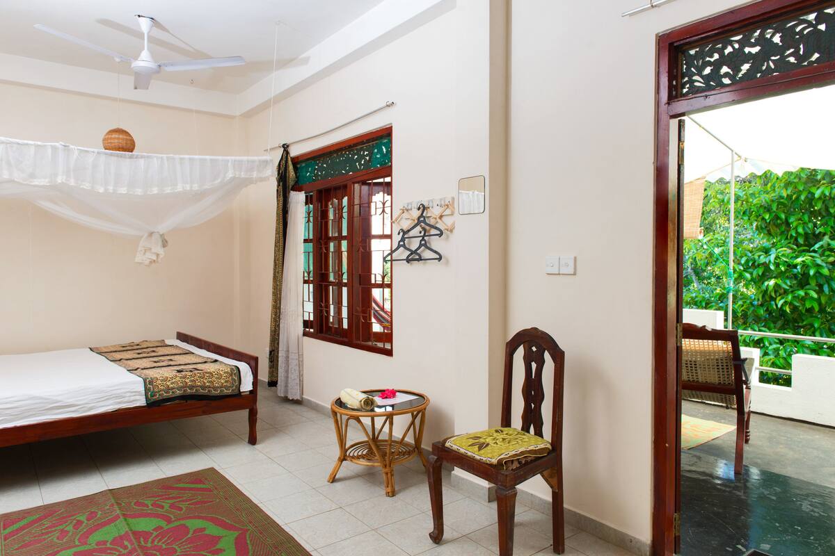 A bedroom with a bed, a chair, a small table, a ceiling fan, and a window with a view of greenery.