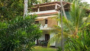 A two-story house with a thatched roof, surrounded by lush greenery and palm trees. - Jungleside House (Hikkaduwa)