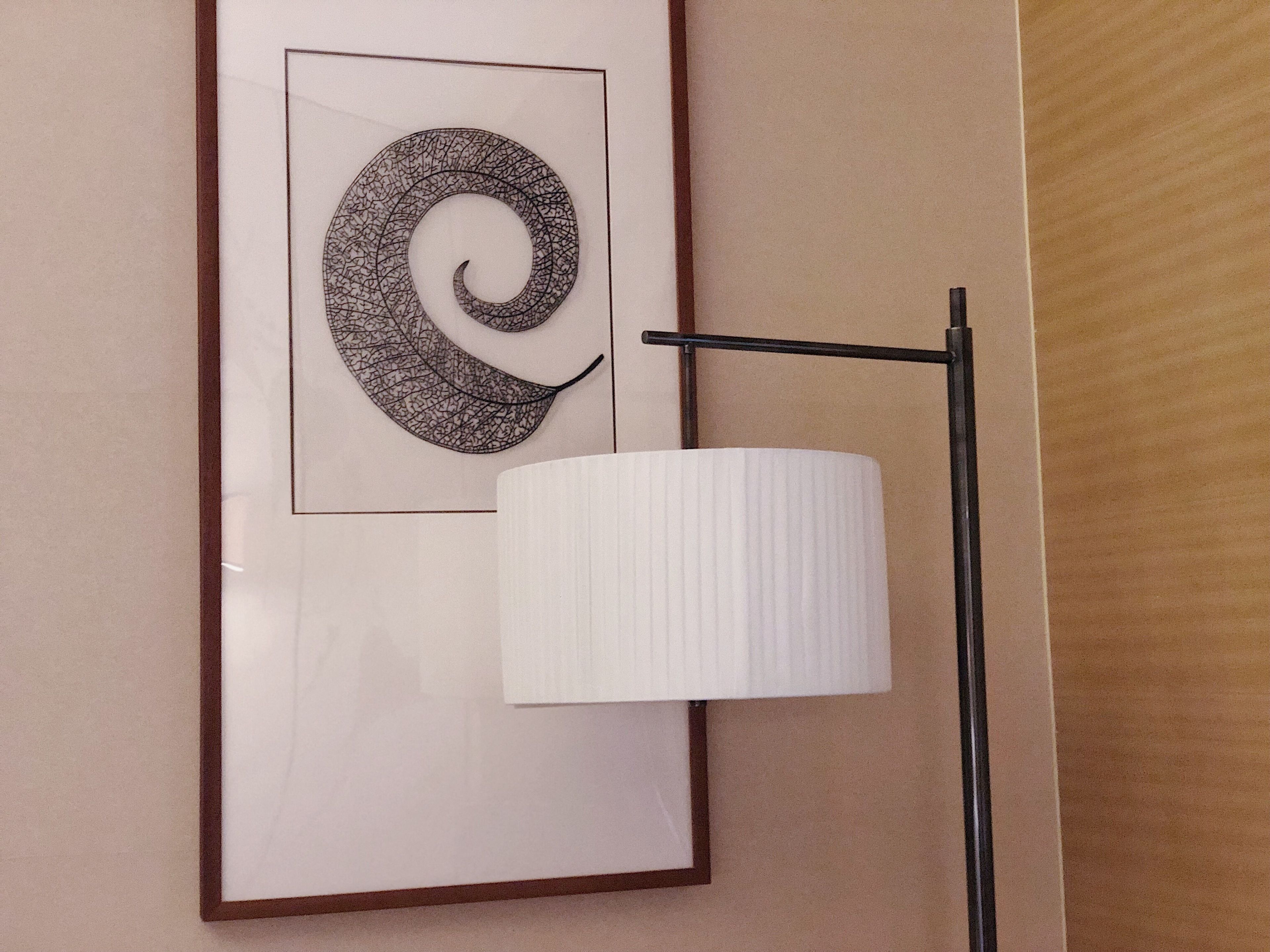A framed abstract artwork with a spiral design on a wall, a white lamp, and a wooden floor.