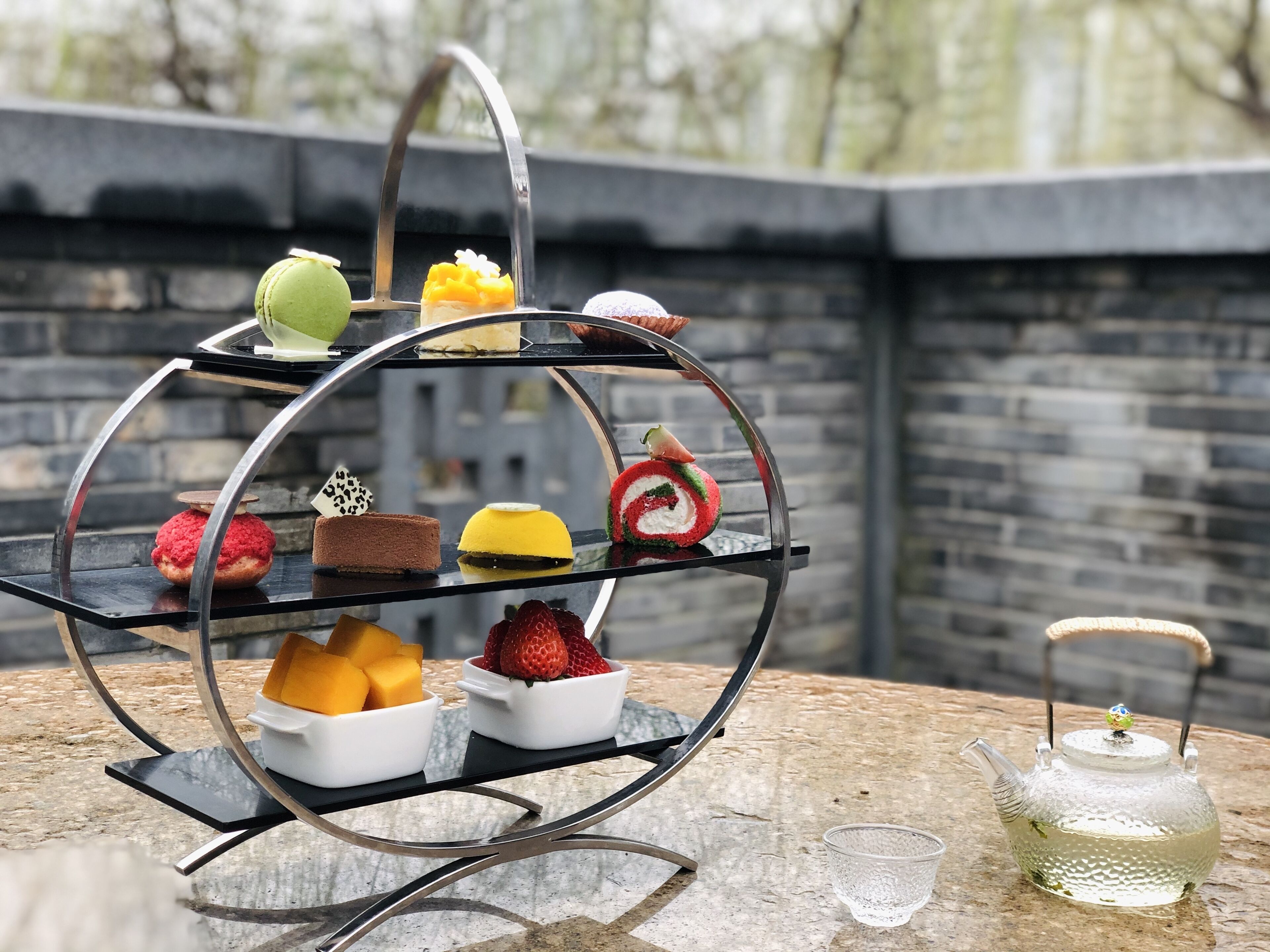 A three-tiered dessert stand with various sweets and a teapot on a stone surface.