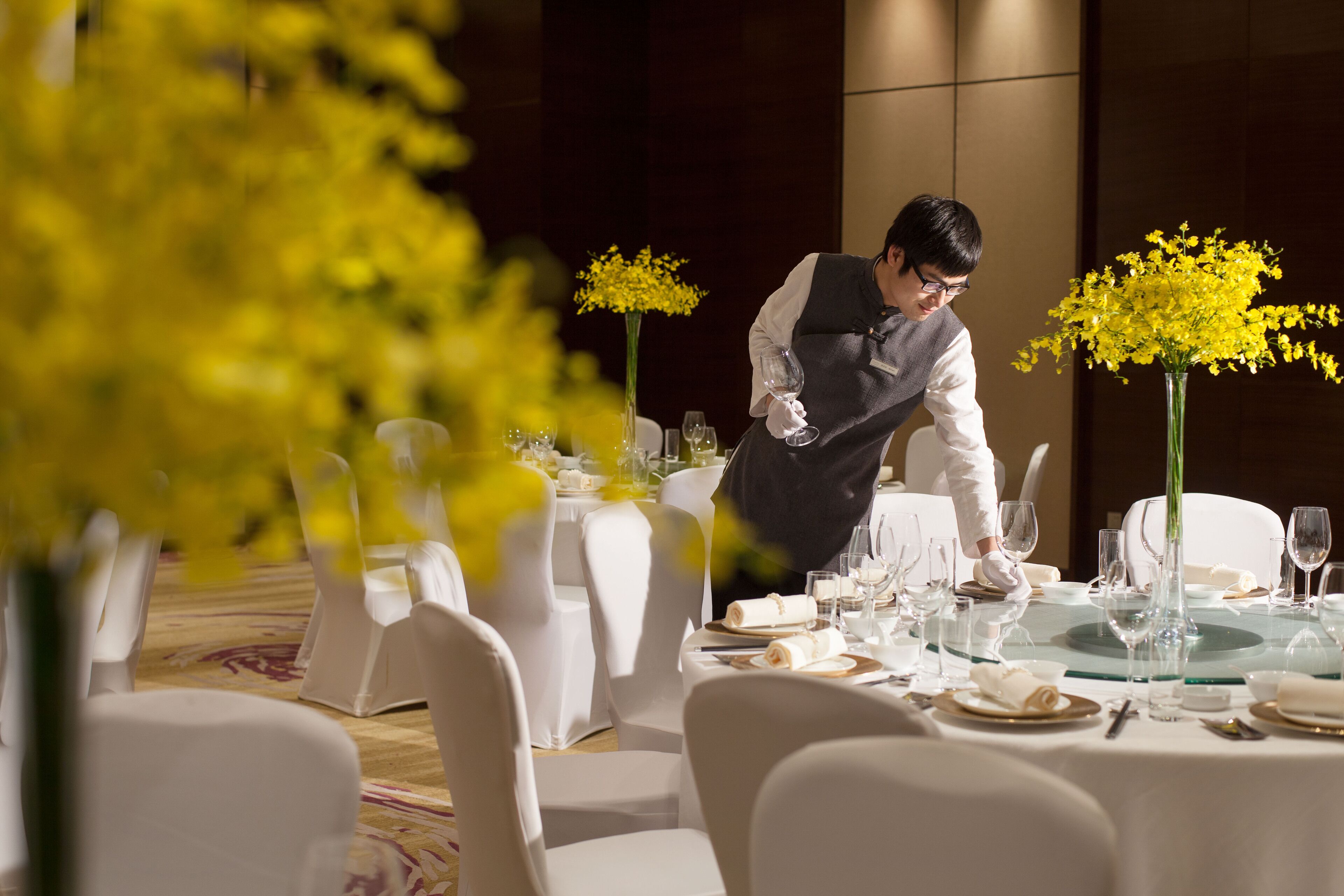 A well-set dining table with white chairs, a waiter attending to the table, and yellow flowers in vases.