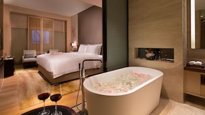 Ein modernes Hotelzimmer mit einem großen Bett, einer mit Rosenblättern gefüllten Badewanne und zwei Weingläsern auf einem Tablett.