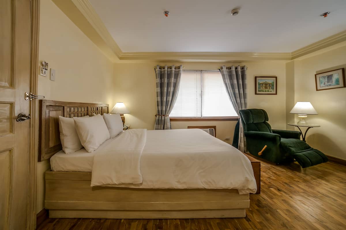 Een hotelkamer met een groot bed, een groene fauteuil, een klein tafeltje met een lamp en een raam met gordijnen.