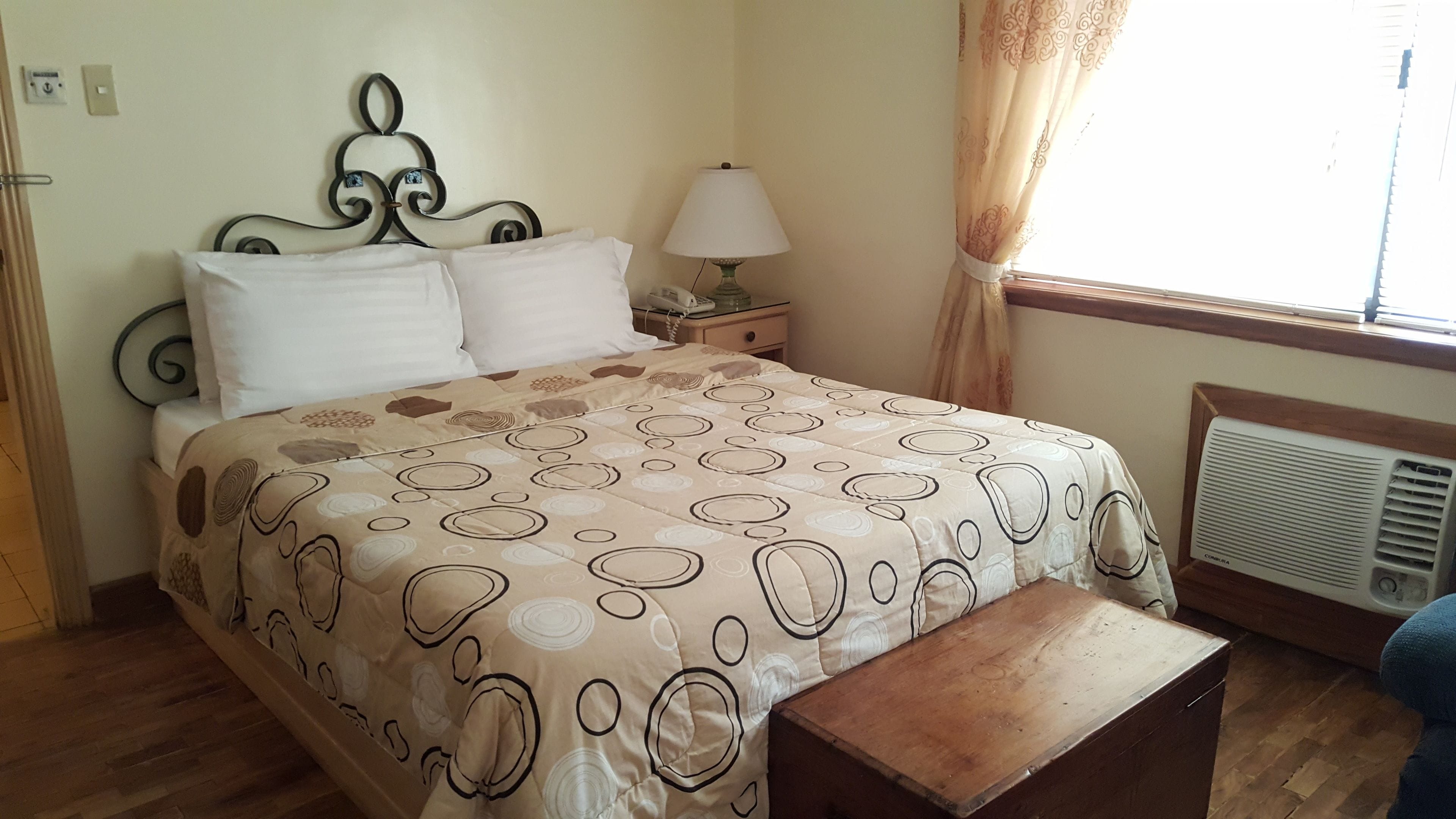 A neatly made bed with a patterned bedspread, a bedside table with a lamp, and a window with curtains.