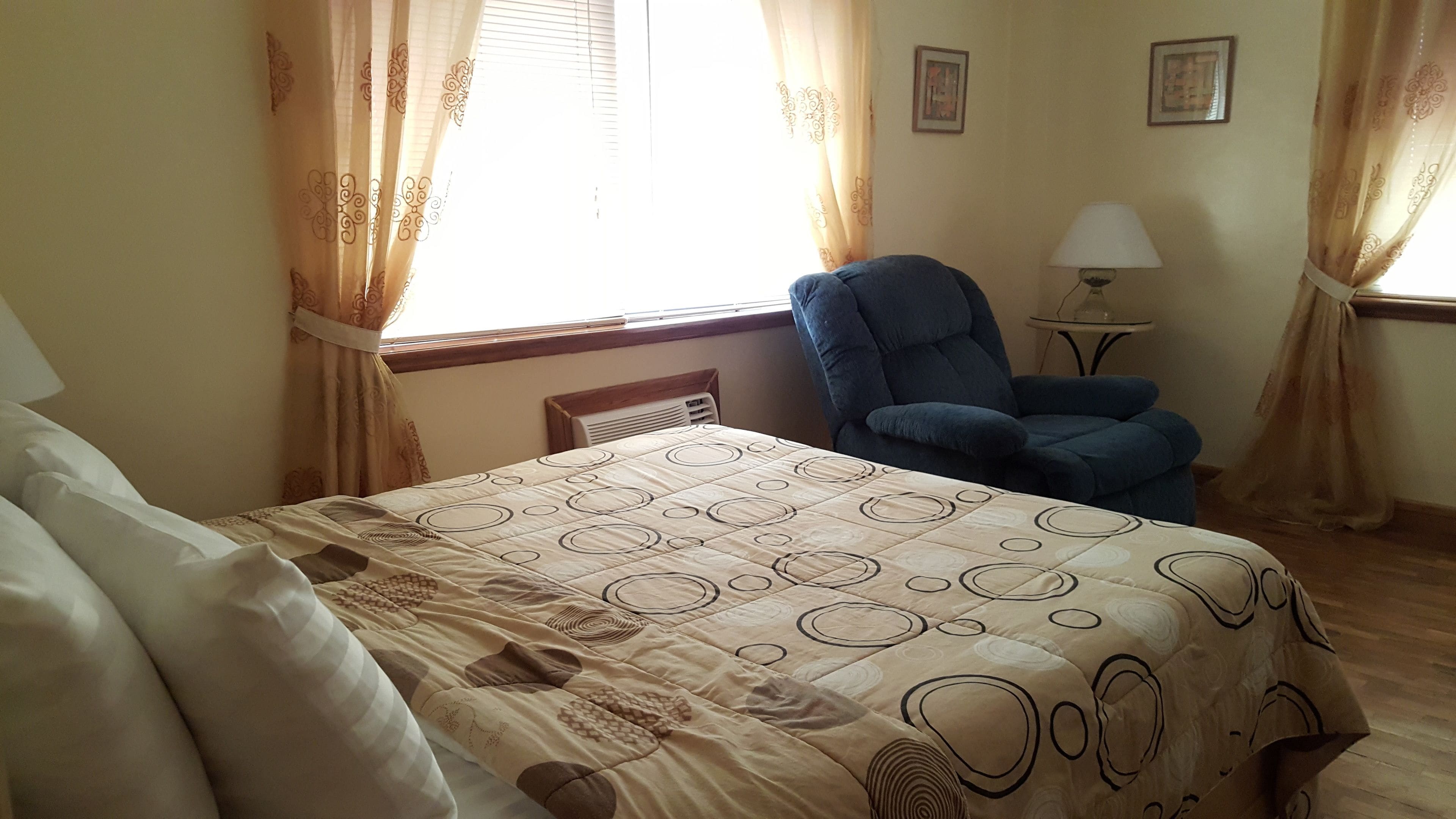 Un dormitorio con cama, una silla, una lámpara y una ventana con cortinas.