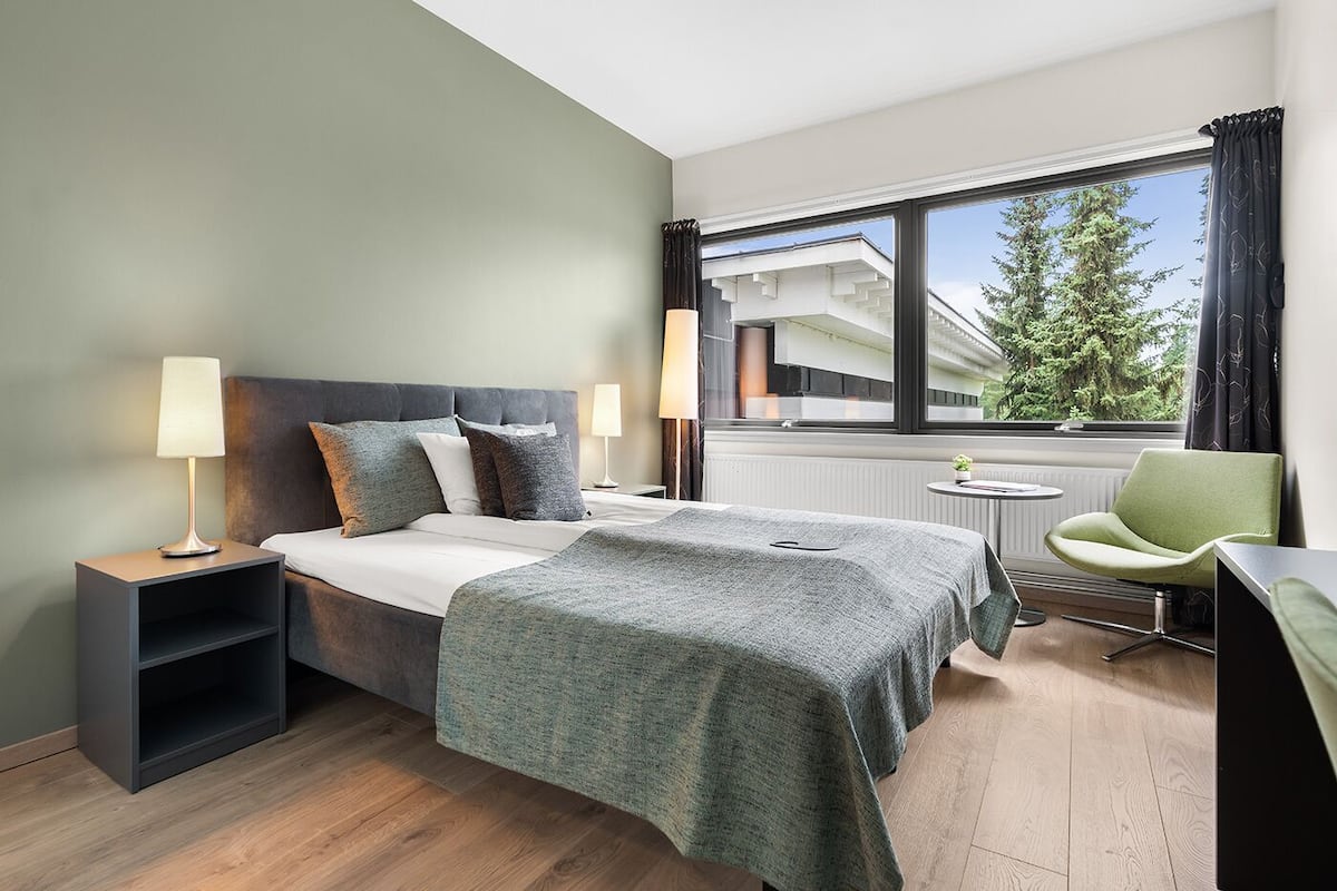 Una habitación de hotel moderna con cama, mesitas de noche, un escritorio y vistas a un edificio y árboles por la ventana.