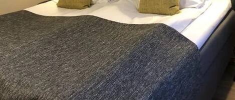 Un lit bien fait, recouvert d’une couverture grise et agrémenté de deux coussins décoratifs, un tableau accroché au mur et deux lampes de chevet.
