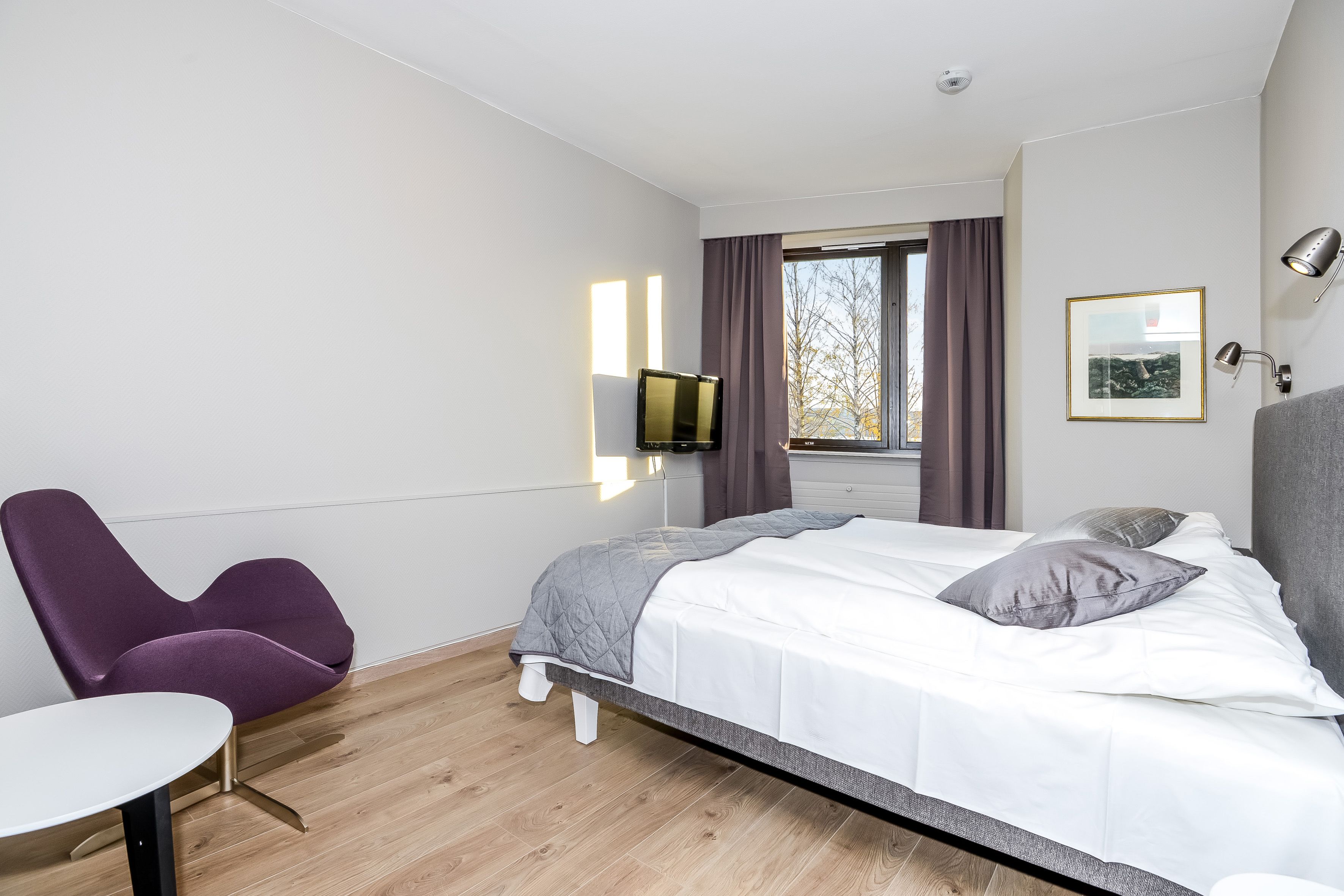 A modern hotel room with a bed, a chair, a small table, a TV, and a window with curtains.