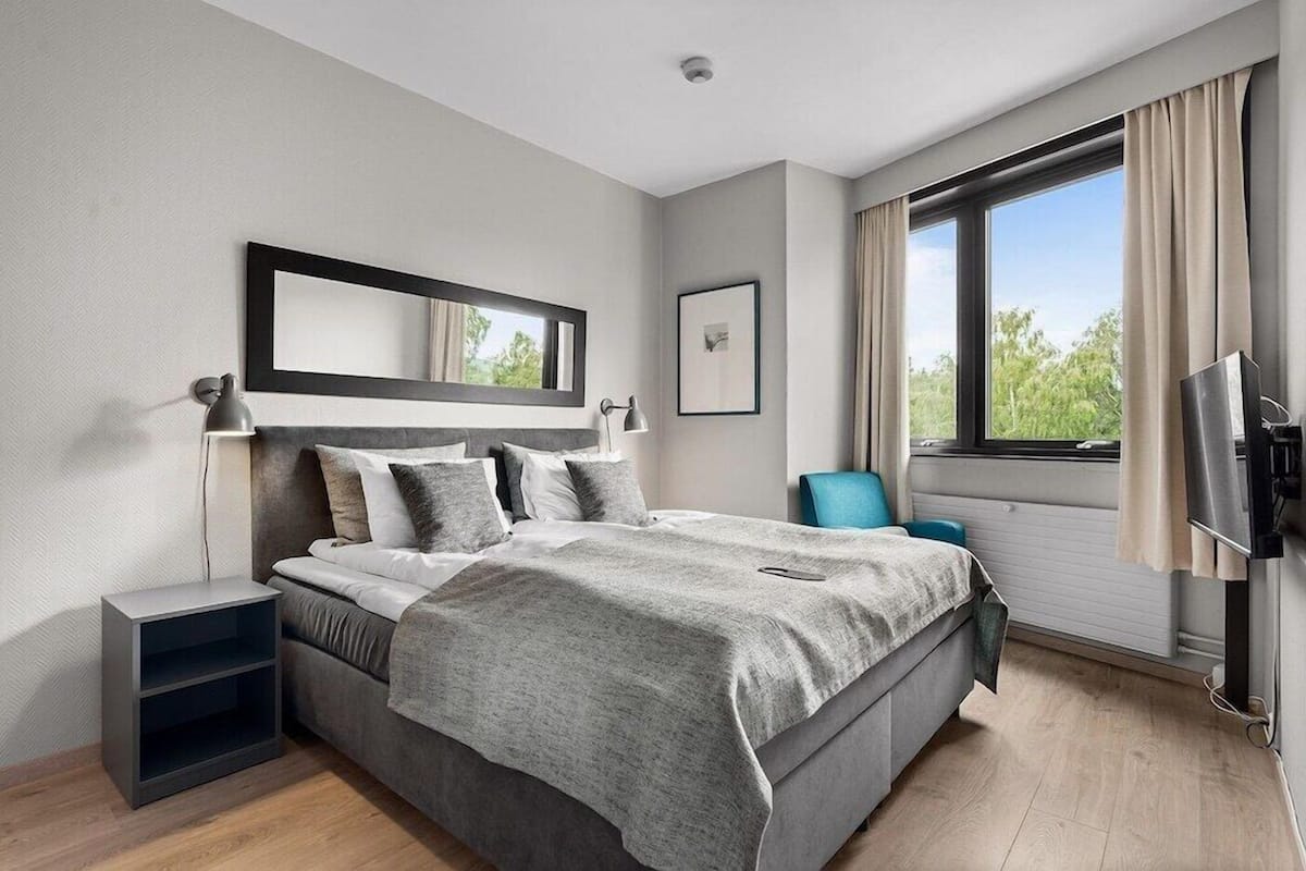 Un dormitorio moderno con cama, mesitas de noche, una silla, televisor y ventana con cortinas.