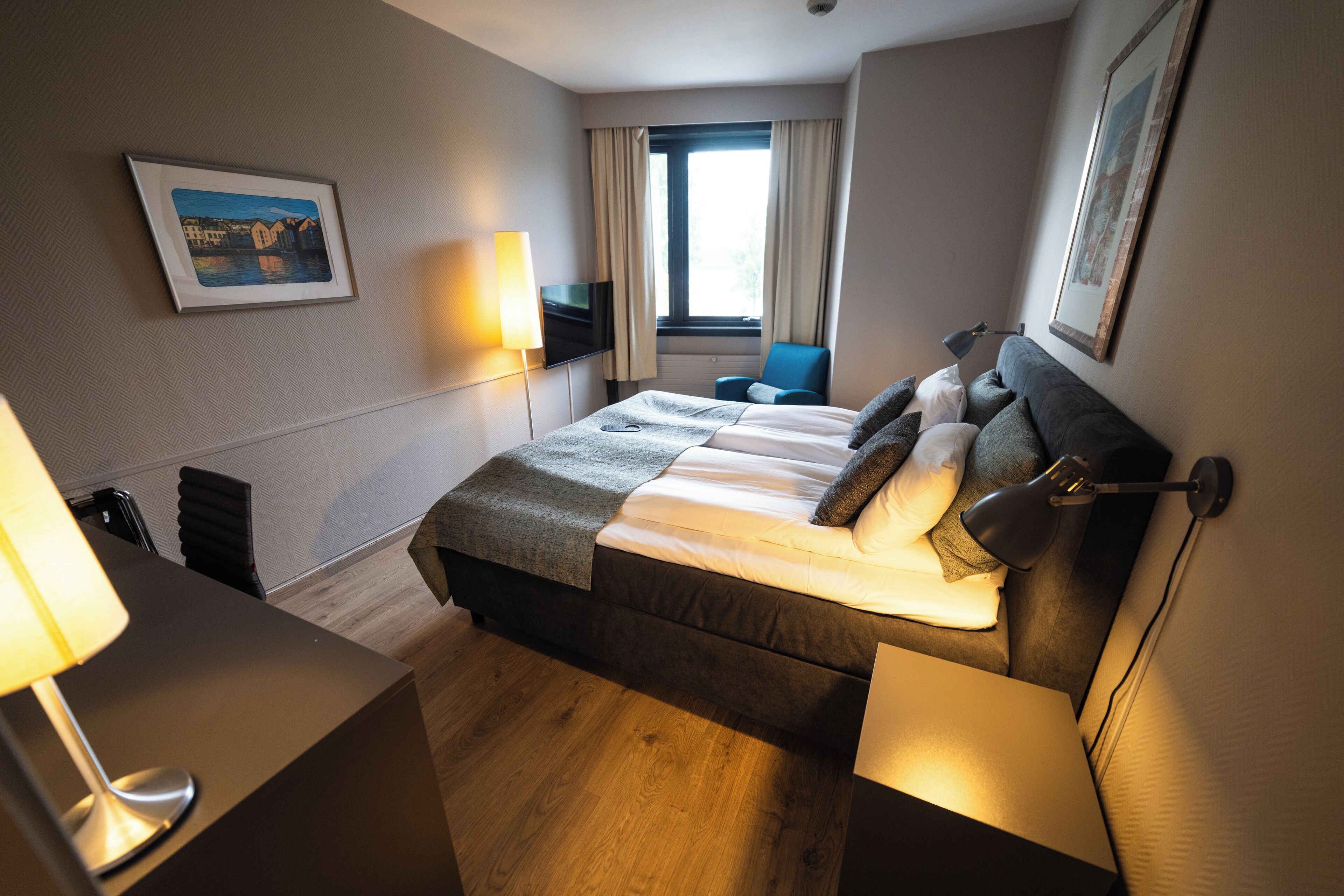A hotel room with a large bed, a desk with a lamp, a chair, a TV, and a window with curtains.