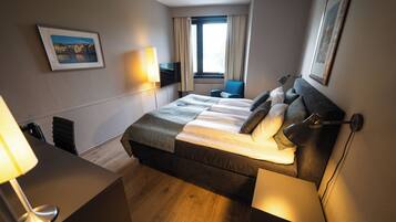 A hotel room with a large bed, a desk with a lamp, a chair, a TV, and a window with curtains.
