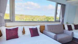 Ein Hotelzimmer mit einem großen Bett, einem grauen Sessel und Blick auf eine grüne Landschaft durch große Fenster.