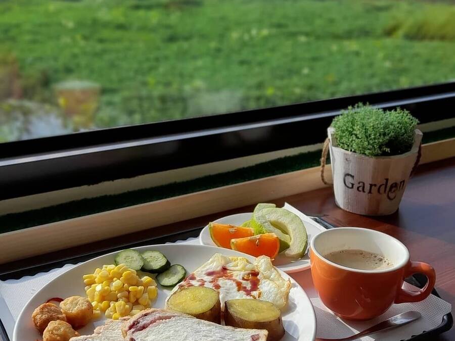 A meal tray with bread, vegetables, corn, and a cup of coffee on a table by a window with a view of greenery.
