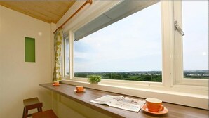 Ein Zimmer mit einem Holztisch, zwei Stühlen, einem Fenster mit Landschaftsblick und einer kleinen Pflanze auf der Fensterbank.