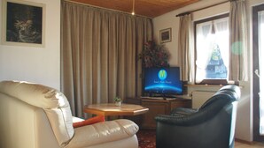 Een woonkamer met een beige bank, een zwarte fauteuil, een houten salontafel en een televisie waarop een afbeelding van de ruimte te zien is.