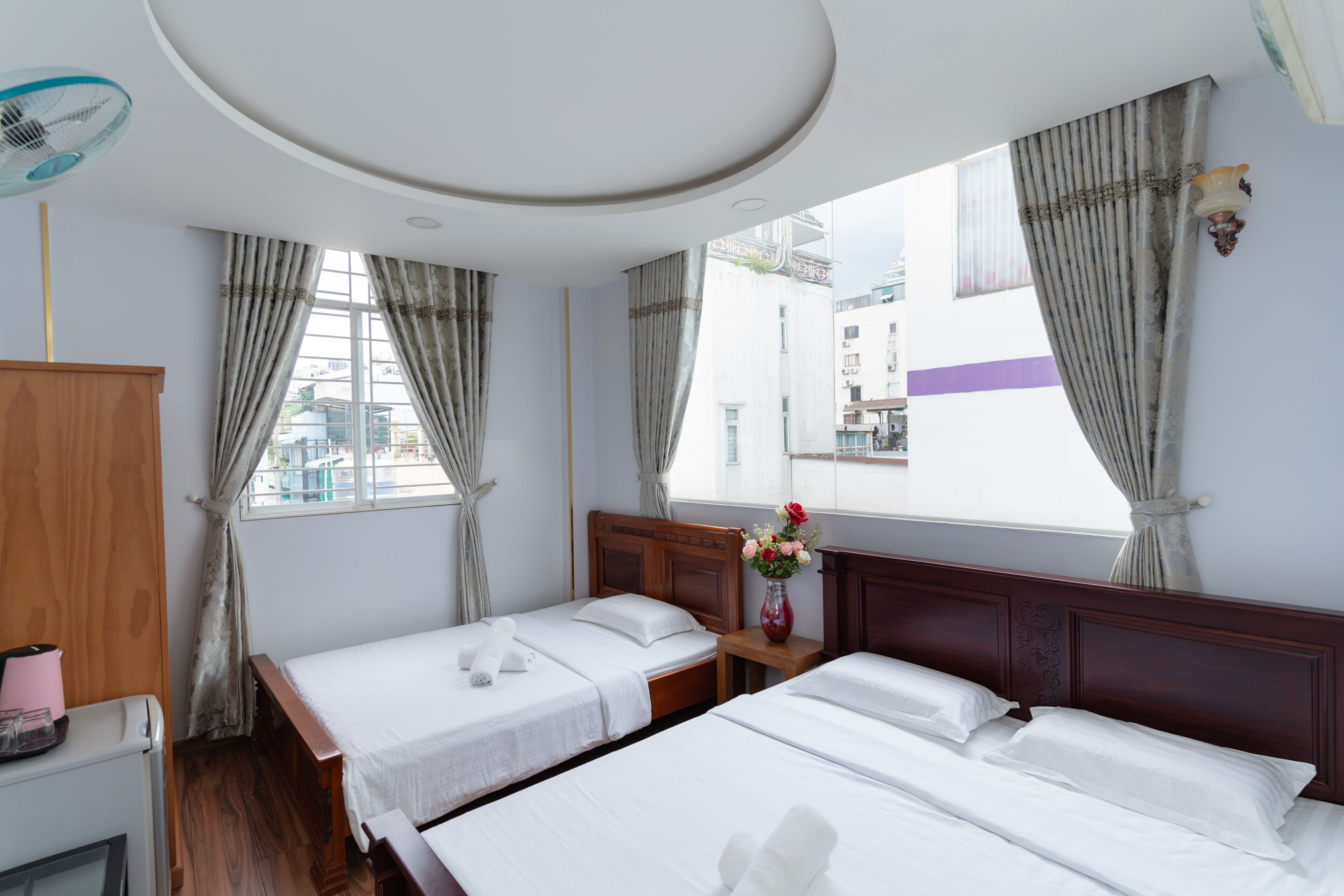 A hotel room with two beds, a wooden headboard, a bedside table with a vase of flowers, and a view of the city through large windows.