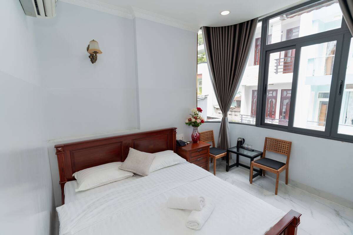 Habitación de hotel con una cama grande, un escritorio, una silla, un florero con flores y una ventana con cortinas.