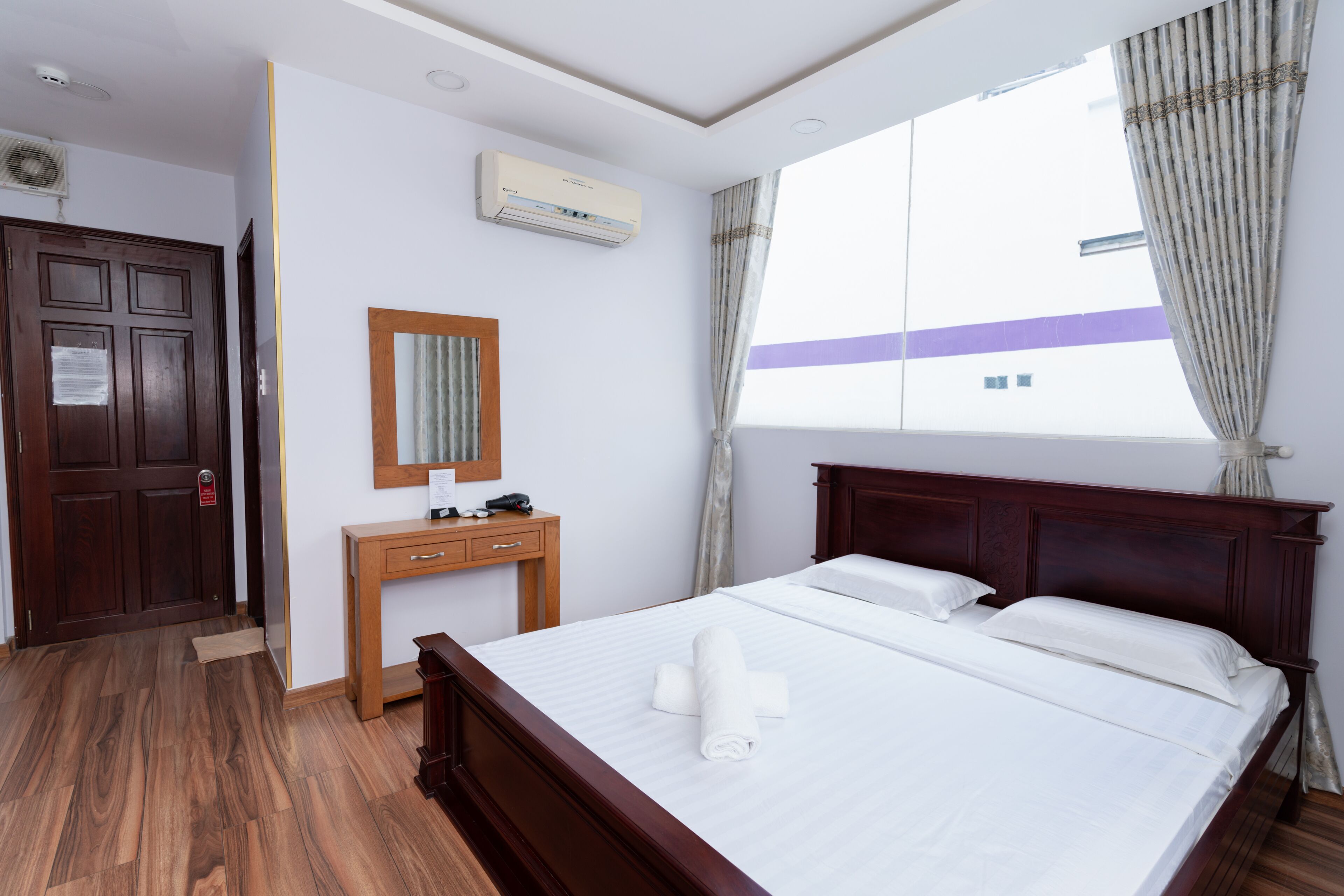 A hotel room with a wooden bed, a wooden desk, a mirror, an air conditioning unit, and a window with curtains.